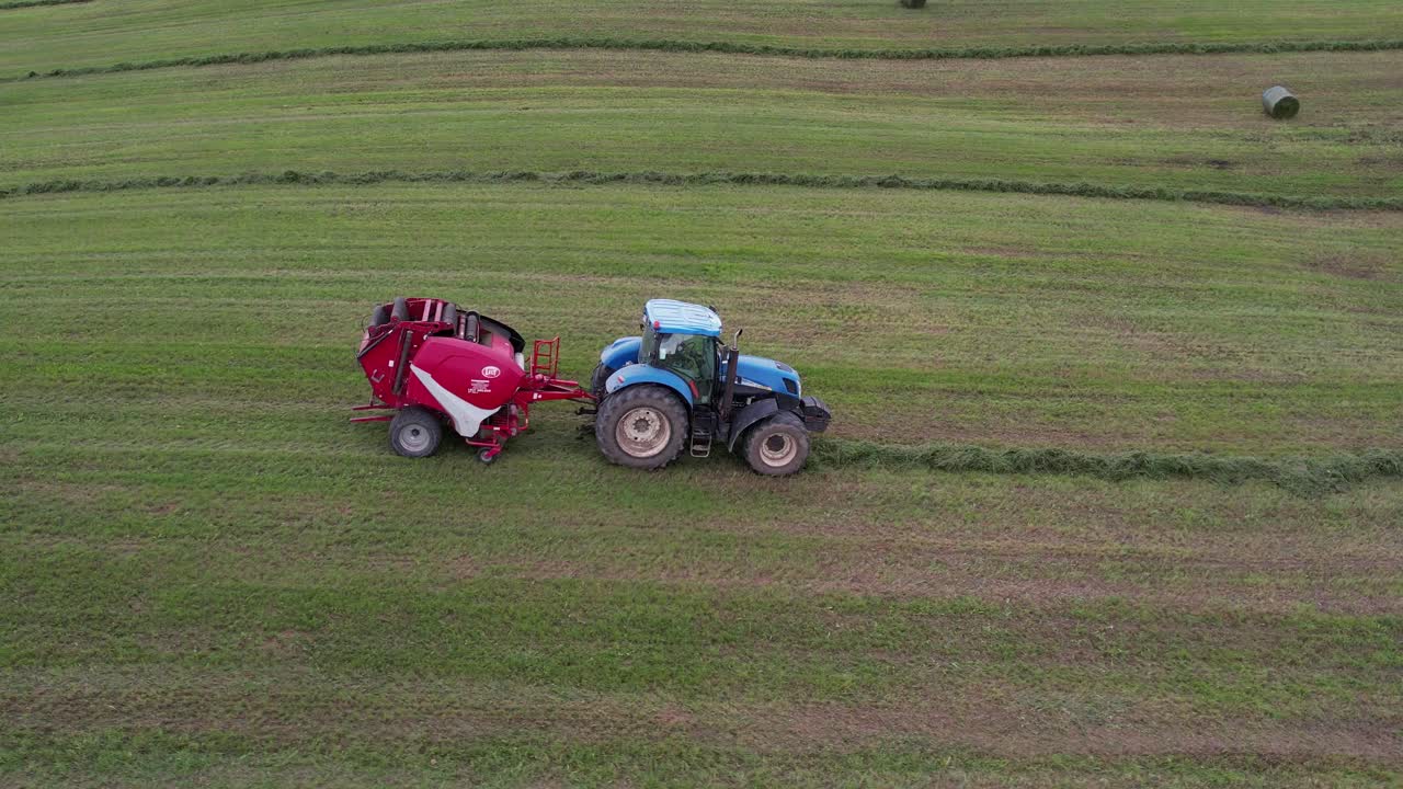 Vista laterale a volo di drone di un trattore blu che traina una rotopressa rossa lungo un andana in un prato verde, con balle di fieno finite sparse in lontananza