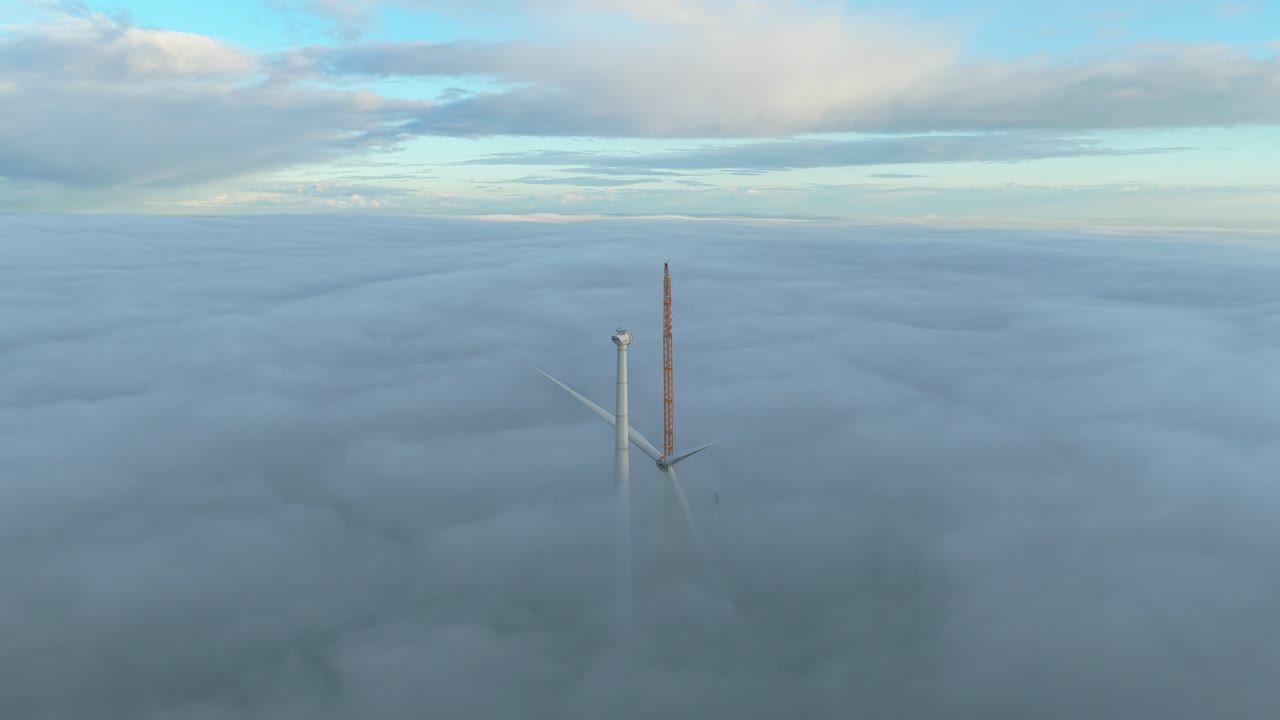 Foggy landscape with an orange crane adding blades to a rising wind turbine