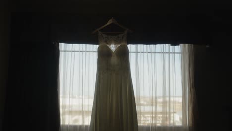 beautiful white wedding dress hanging on clothes hanger, backlit curtains, moody scenic push in movement in cinematic slowmotion shot