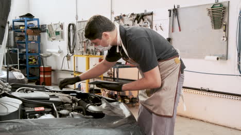 Hispanic-male-car-mechanic-wearing-mask,-holding-clipboard,-checking-under-hood