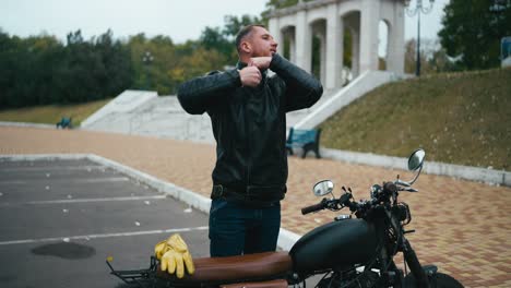 man in leather jacket standing beside a classic motorcycle in a park