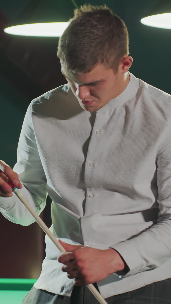 hombre de juego con camisa blanca y pantalones grises se sienta en la mesa de billar verde, sosteniendo el palo de la pilla y aplicando cuidadosamente la cubierta roja con expresión enfocada a medida que la cámara hace zoom.