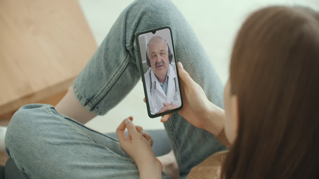 Beautiful woman checks possible symptoms with professional physician using online video chat. Young girl sick at home using smartphone to talk to her doctor via video conference medical app.