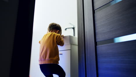 Small Caucasian kid stands on the prop near the sink. Toddler stepping quickly and laughing cheerfully. Low angle view.