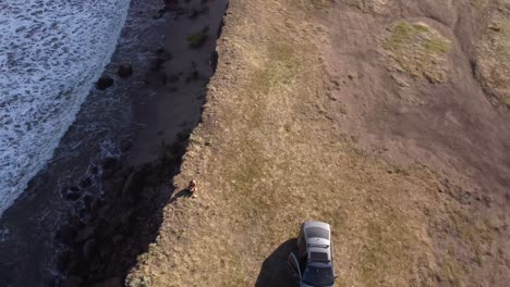 Aerial-top-down-of-lonely-woman-parking-with-car-close-to-steep-cliffs-edge-and-enjoying-ocean-view-during-sunset