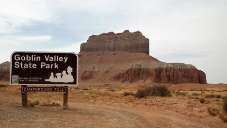 Tiro-Inclinado-Hacia-Arriba-Que-Revela-El-Cartel-Del-Parque-Estatal-Goblin-Valley-Con-Una-Gran-Roca-Roja-En-Medio-Del-Seco-Desierto-De-Utah-En-Un-Cálido-Día-Soleado-De-Verano