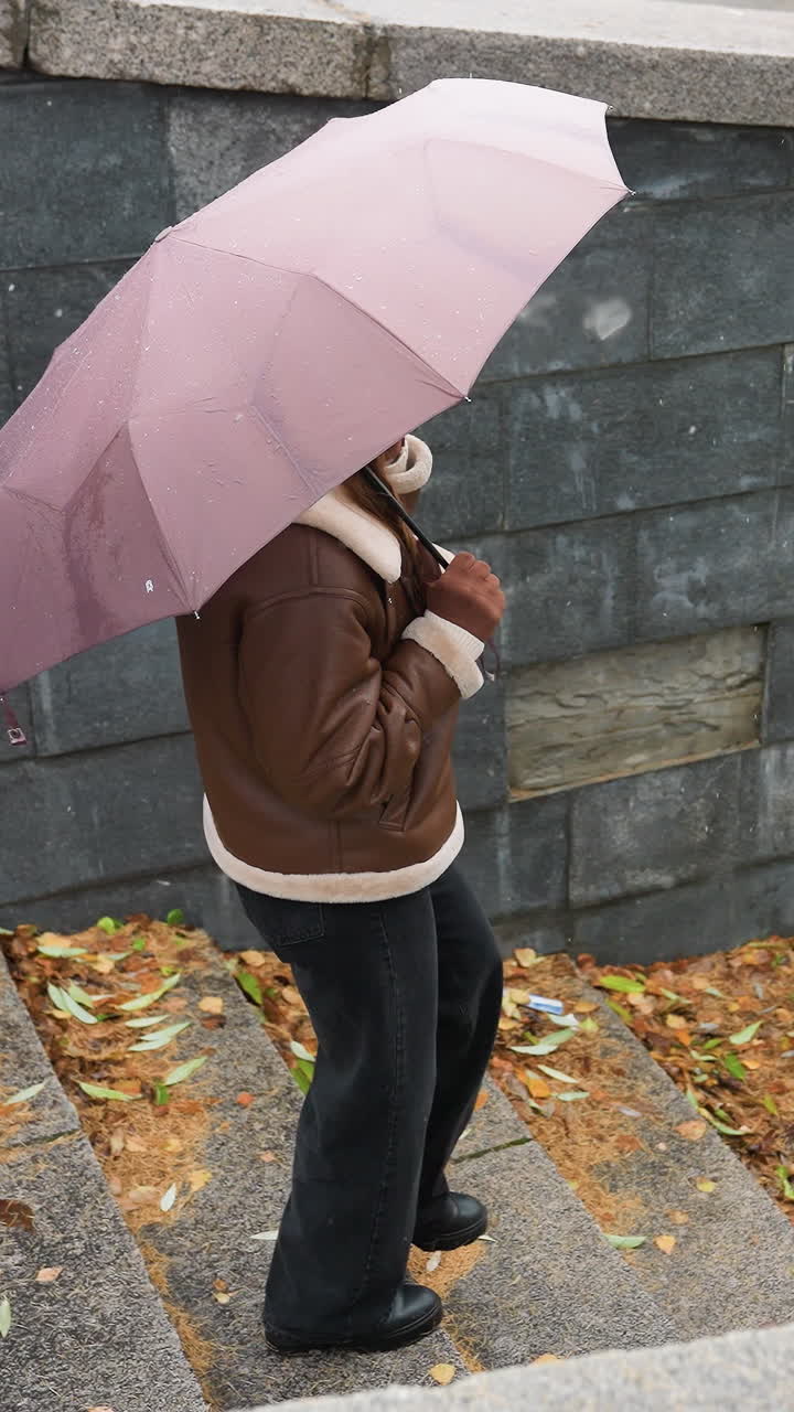 Up shot of young girl smiling on phone call holding umbrella, wearing knit cap, brown shearling jacket, black trousers, walking down stone steps during light snowfall with colorful autumn leaves