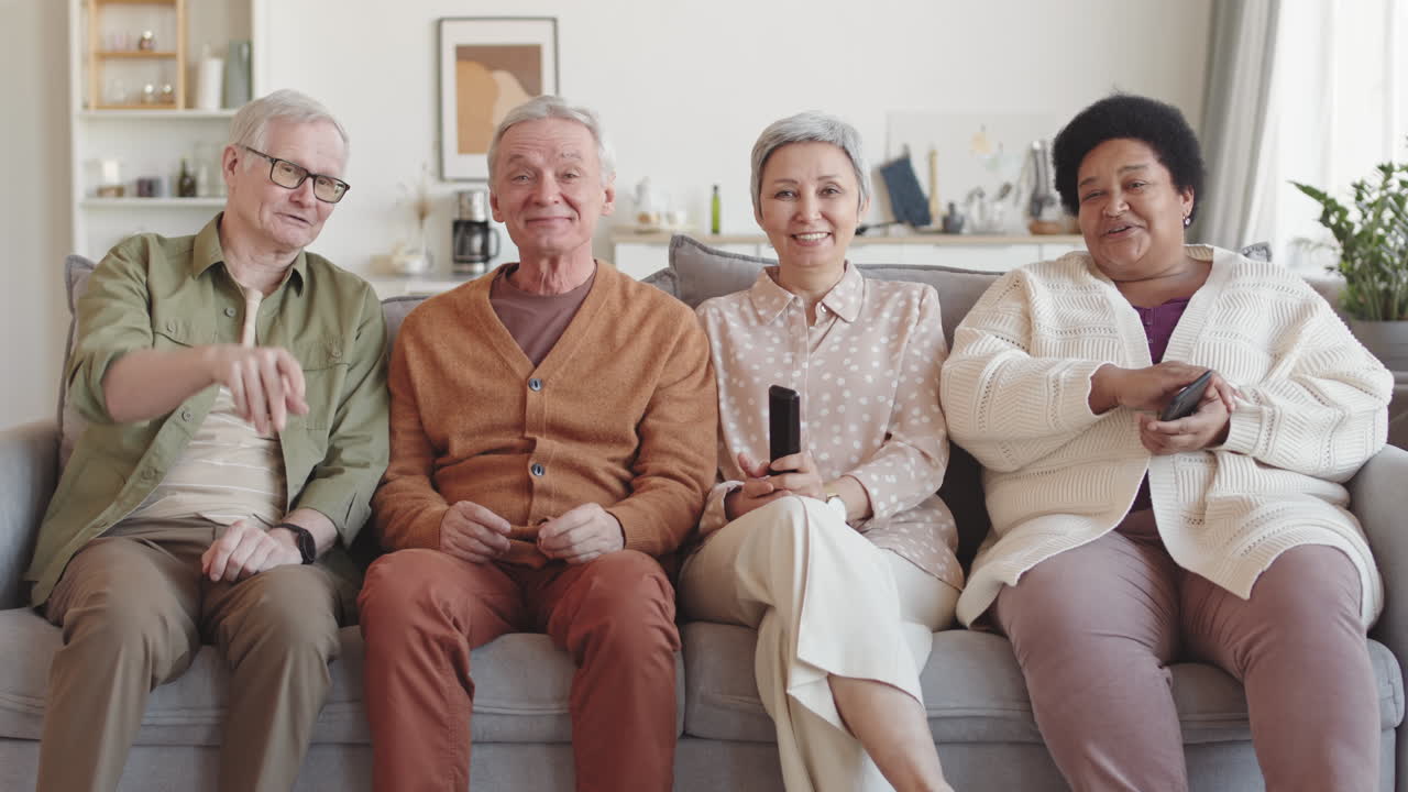 Medium long of four multiethnic diverse senior friends sitting together on sofa in living room, watching invisible TV, pointing fingers forward, talking and laughing