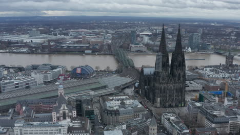 Aerial-view-of-city-along-wide-Rhine-river-with-muddy-water.-Majestic-gothic-Cologne-Cathedral-with-tall-towers-and-Main-train-station.-Cologne,-Germany