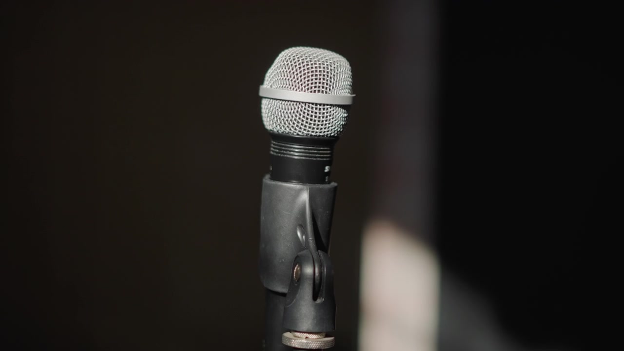 Close up microphone on dark stage, empty rehearsal space with spotlight glow, worn metal mesh and cable, sturdy stand, intimate coaching vibe, gritty texture, low light mood, performer preparation