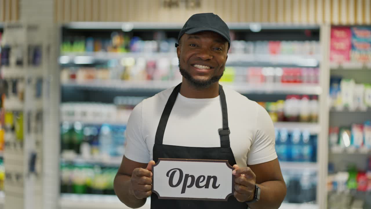 A happy man with Black skin color in a white T-shirt and a black apron stands near the counters of a large modern grocery supermarket and holds a sign that says open