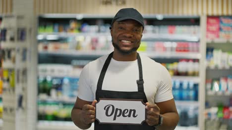 A-happy-man-with-Black-skin-color-in-a-white-T-shirt-and-a-black-apron-stands-near-the-counters-of-a-large-modern-grocery-supermarket-and-holds-a-sign-that-says-open