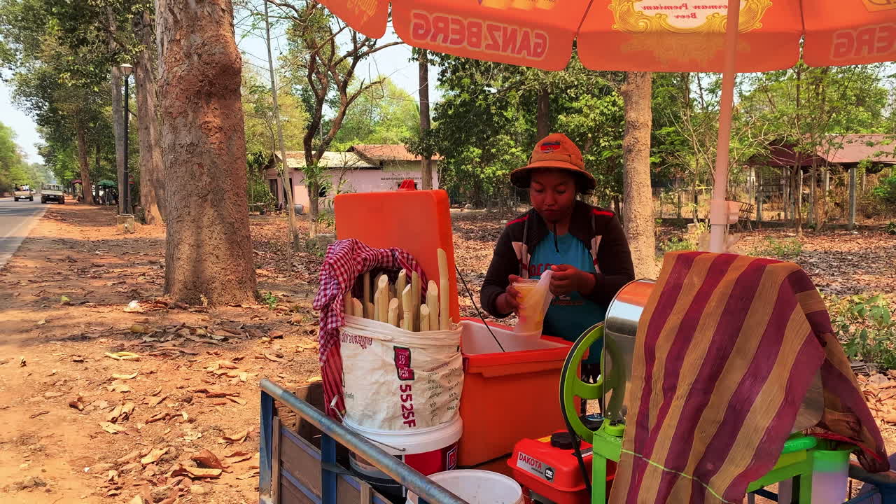 A Cambodian woman pours sugarcane juice into a plastic cup from a cart at the side of the road, wide shot