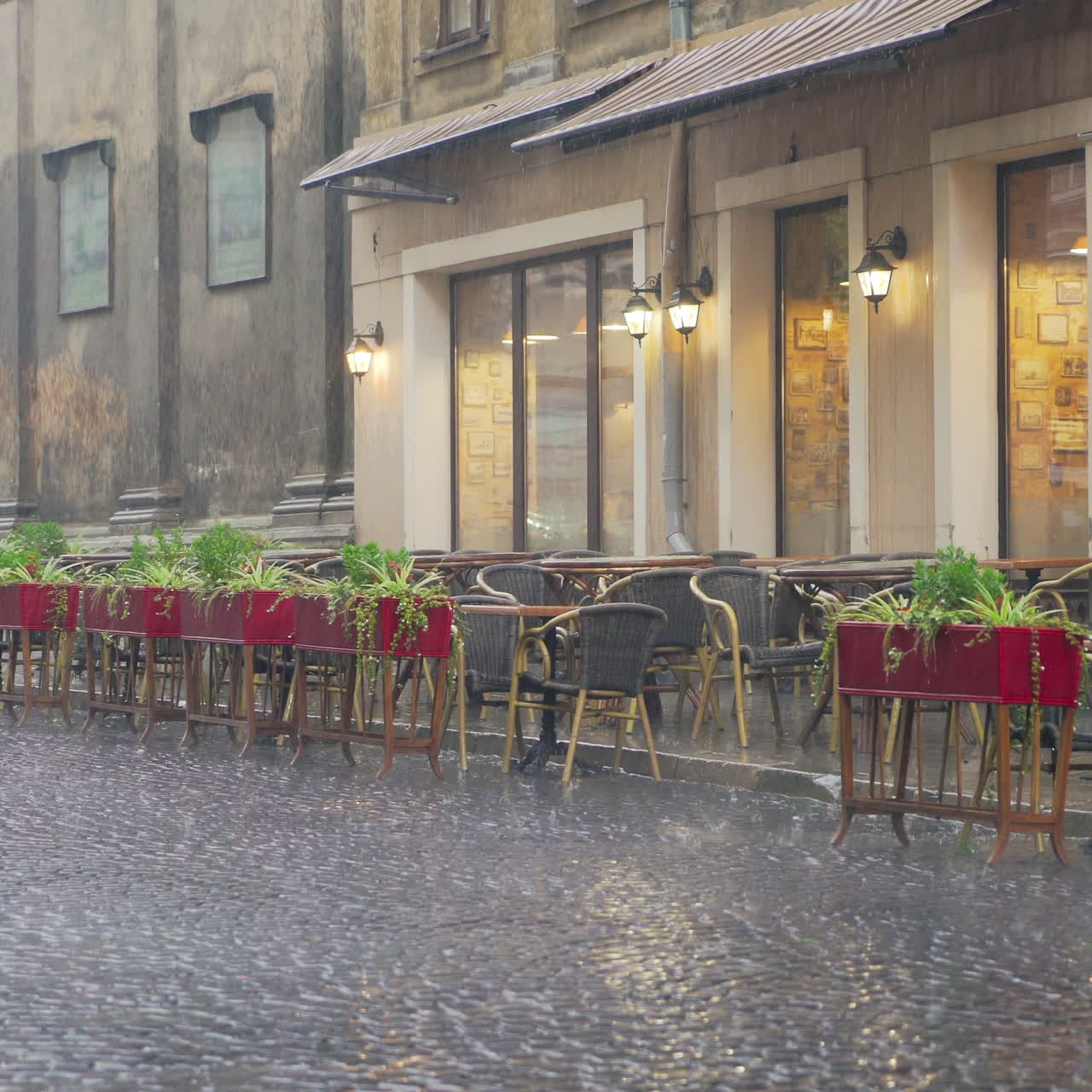 Heavy raindrops falling outside on the cafe's table and chairs in the evening. Big drops of rain are falling on the pavement and flower pots on the background of the empty restaurant outdoors