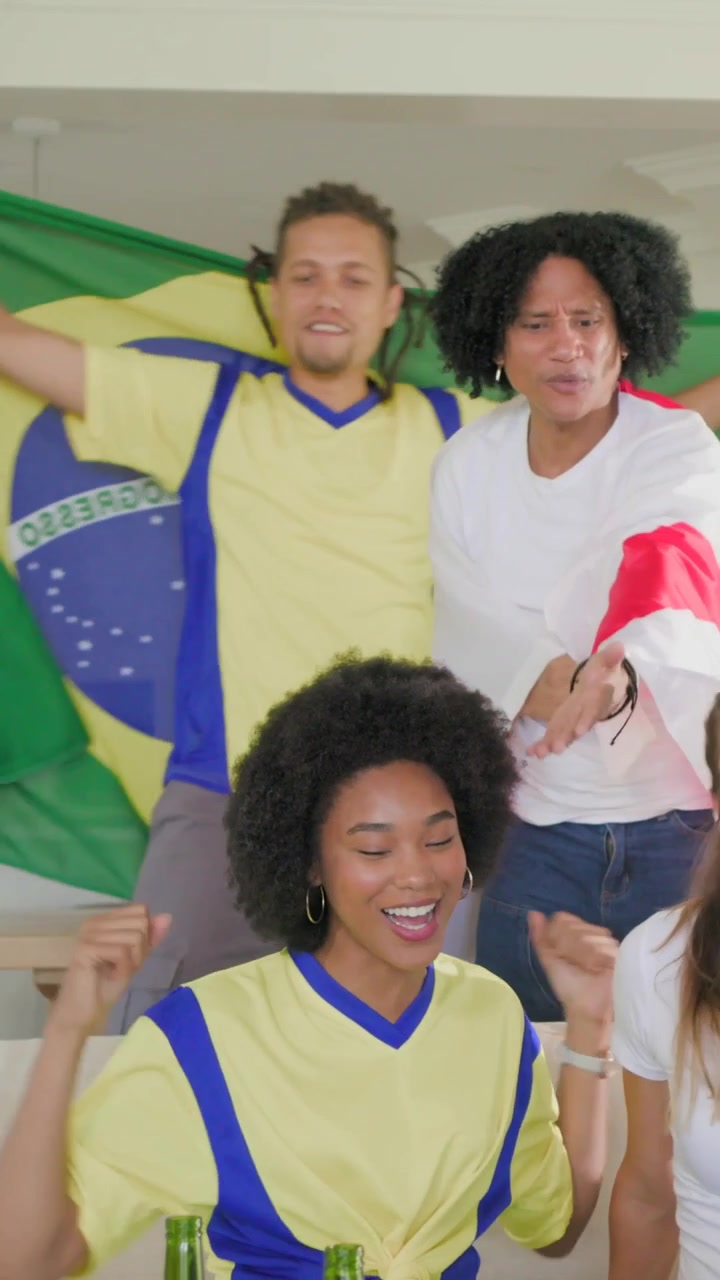 Vídeo vertical: Amigos comemorando após um gol em casa, camisa amarela e bandeiras da Inglaterra e do Brasil tremulando.