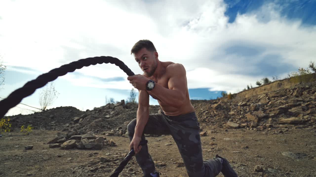 Cross power training. Concentrated sportsman exercises outdoors. Young shirtless muscular man training with battle ropes on rocky background. Slow Motion