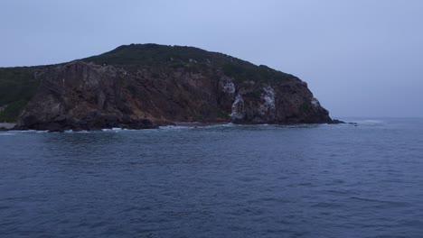 a dramatic seaside cliff stands against the calm pacific ocean as waves gently brush the rugged shoreline under a soft blue sky.