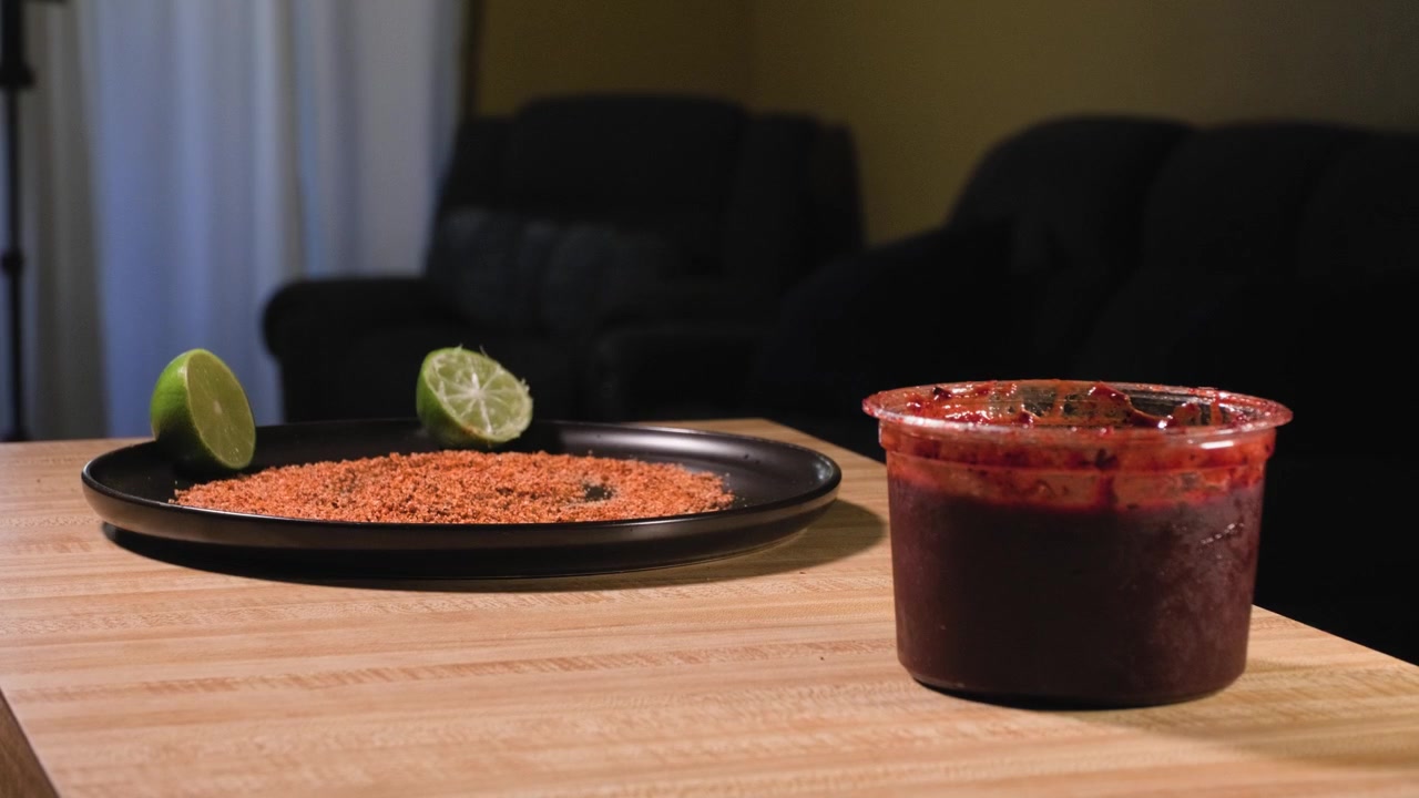 Un hombre toma un vaso de plástico esmerilado con salsa chamoy oscura y lo retira de una mesa de cocina junto a ingredientes frescos