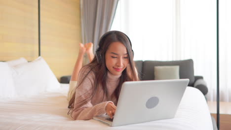 slow-motion of relaxed asian woman using her laptop computer and listening to the music lying on the bed with her legs raised at home wearing casual clothes