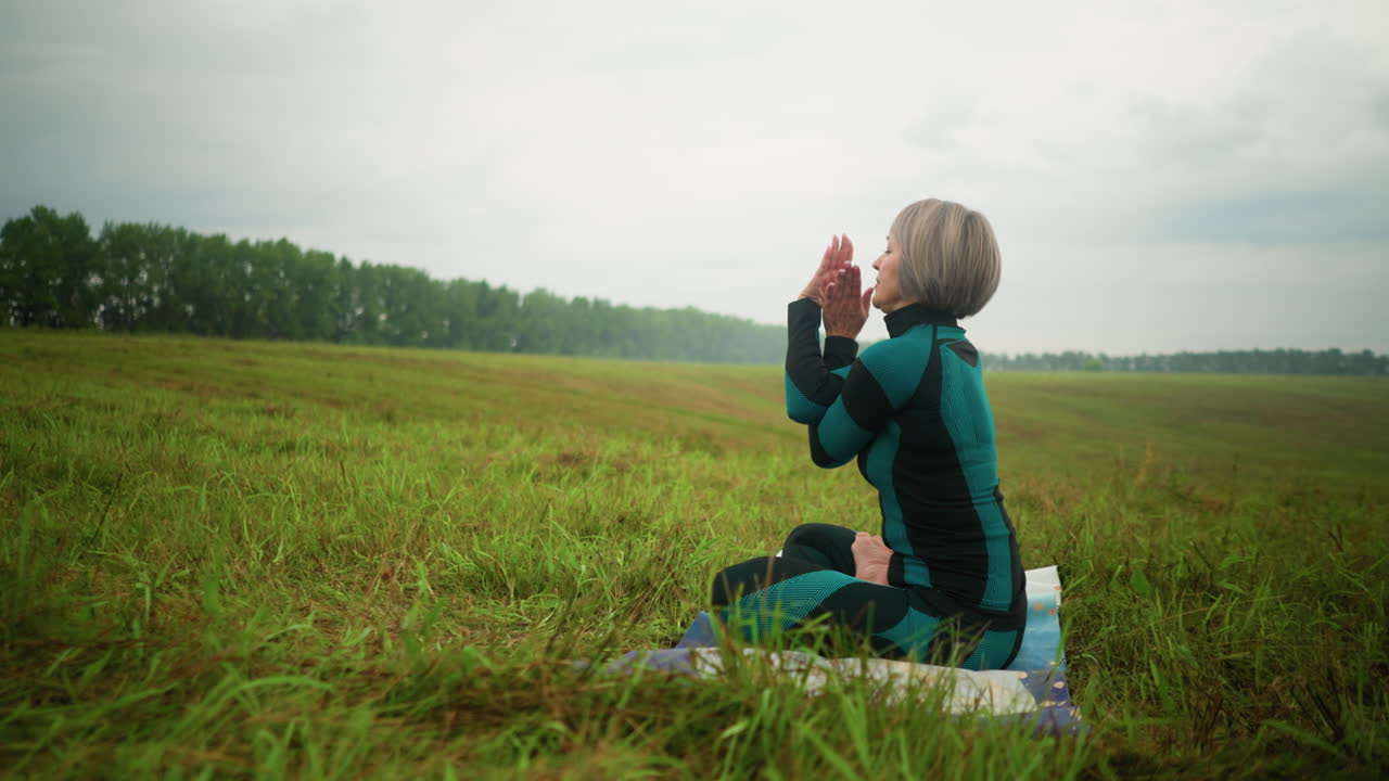 안개가 가득한 에서 요가 매트에 앉아 요가를 연습하는 여성의 측면 뷰, 소 얼굴 요가, 눈이 닫힌, 흐릿한 배경으로 넓은 잔디에서 구름이 가득한 하늘 아래