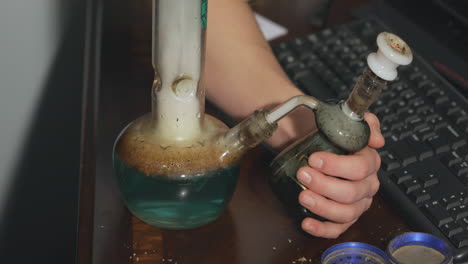 a hand holds and removes an ash catcher as a bowl of marijuana is smoked on an office desk