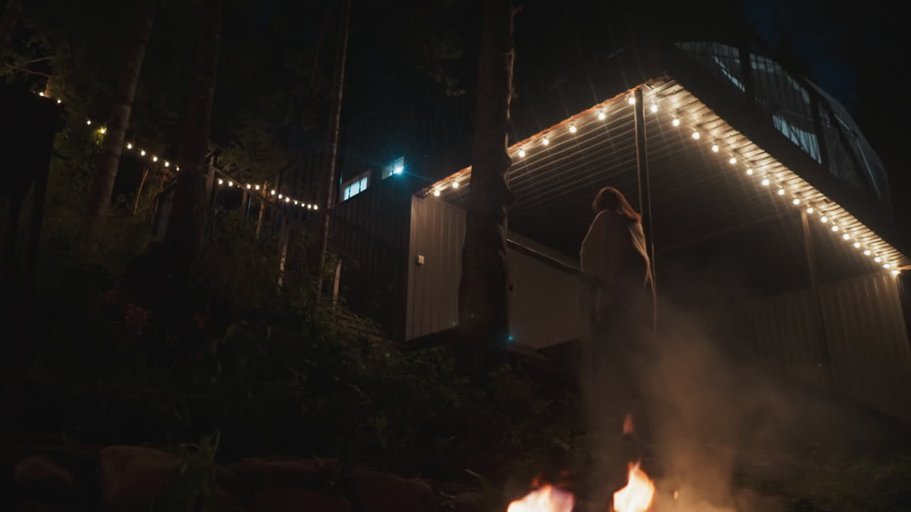 마당에서 계단을 오르는 여자 - 은 숲 사이에서 관광객을위한 시골 집의 사적인 영토의 야간 풍경 - 밤에는 캠프에서 불꽃과 함께 산책합니다.