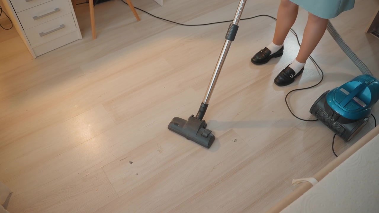 Mulher aspirando chão de madeira clara em quarto iluminado, aspirador de pó tipo canister turquesa com cabo visível puxa ao longo da borda enquanto pés em sapatos pretos se movem pelas tábuas, close-up do bico deslizando, poeira.