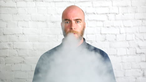 portrait of a man vaping indoors in front of a white wall looking directly at the camera with cloud of smoke