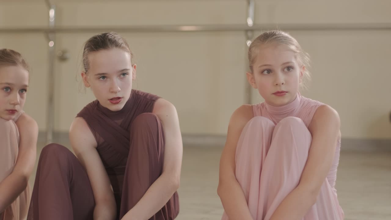 검은색 댄스웨어를 입은 젊은 발레 학생들이 나무 바닥과 벽에 장착된 바를 가진 넓은 발레 스튜디오에서 자세를 연습하는 그룹.