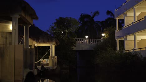 Impresionante-Y-Tranquila-Escena-Nocturna-De-Pequeños-Bungalows-Tropicales-A-Lo-Largo-De-Un-Pequeño-Río-Con-Un-Puente-Iluminado-Arriba-En-Un-Hermoso-Y-Exótico-Complejo-Vacacional-En-Riviera-Maya,-México,-Cerca-De-Cancún-Y-Tulum