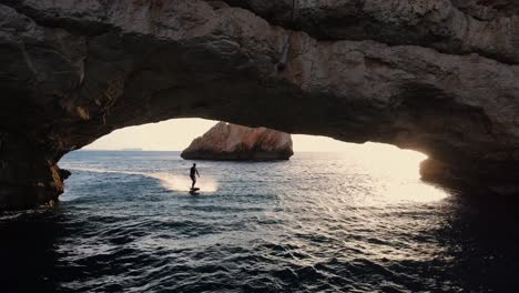 silhouette of an electric surfboard rider cruising through a rock archway in the water