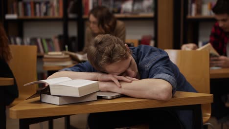 Young-European-Student-In-A-College-Library-Or-Class-Sleeping-On-Desk-With-Pile-Of-Books-On-It