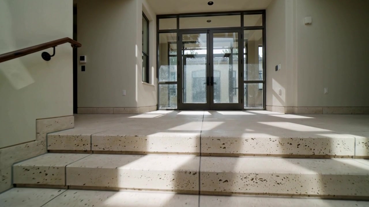Câmera em movimento revelando degraus de pedra salpicados e porta de metal e vidro no hall de entrada, com a luz solar se deslocando.