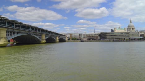 POV-view-of-the-London-Thames-standing-by-the-fence-many-iconic-historical-buildings-Canary-Wharf-in-the-background-sunny-day-with-clouds-birds-flying-moving-motion-to-the-side-cinematic