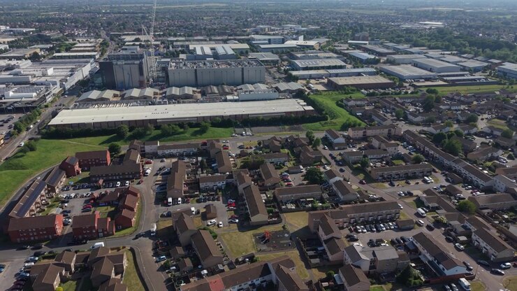 Drone captures an expansive aerial view of a massive data center complex under construction, adjacent to a sunny residential neighborhood with houses and parked cars.