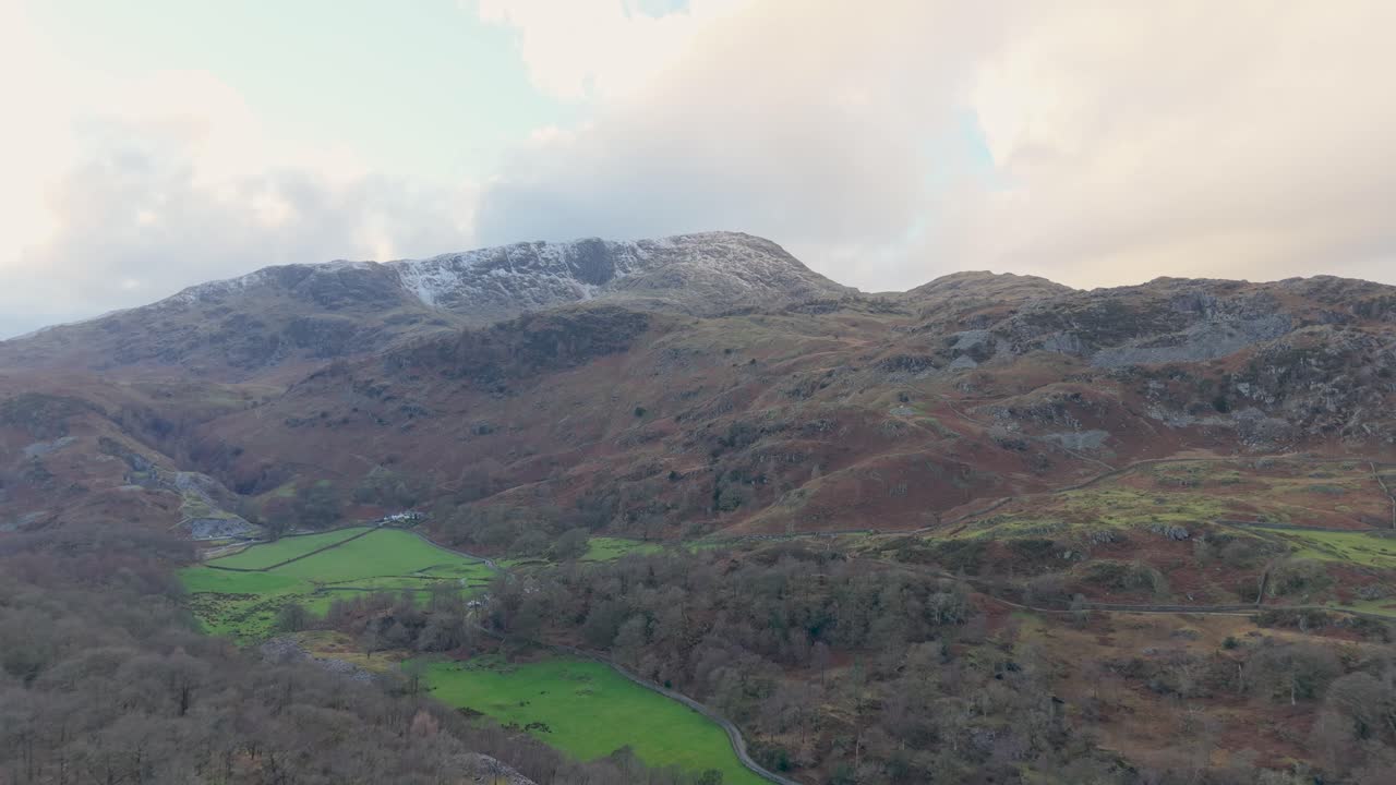 Forward drone flight toward a distant mountain with a small snowy peak. Green and brown terrain rises in the beneath large white clouds and a slightly gloomy sky, creating a moody atmosphere