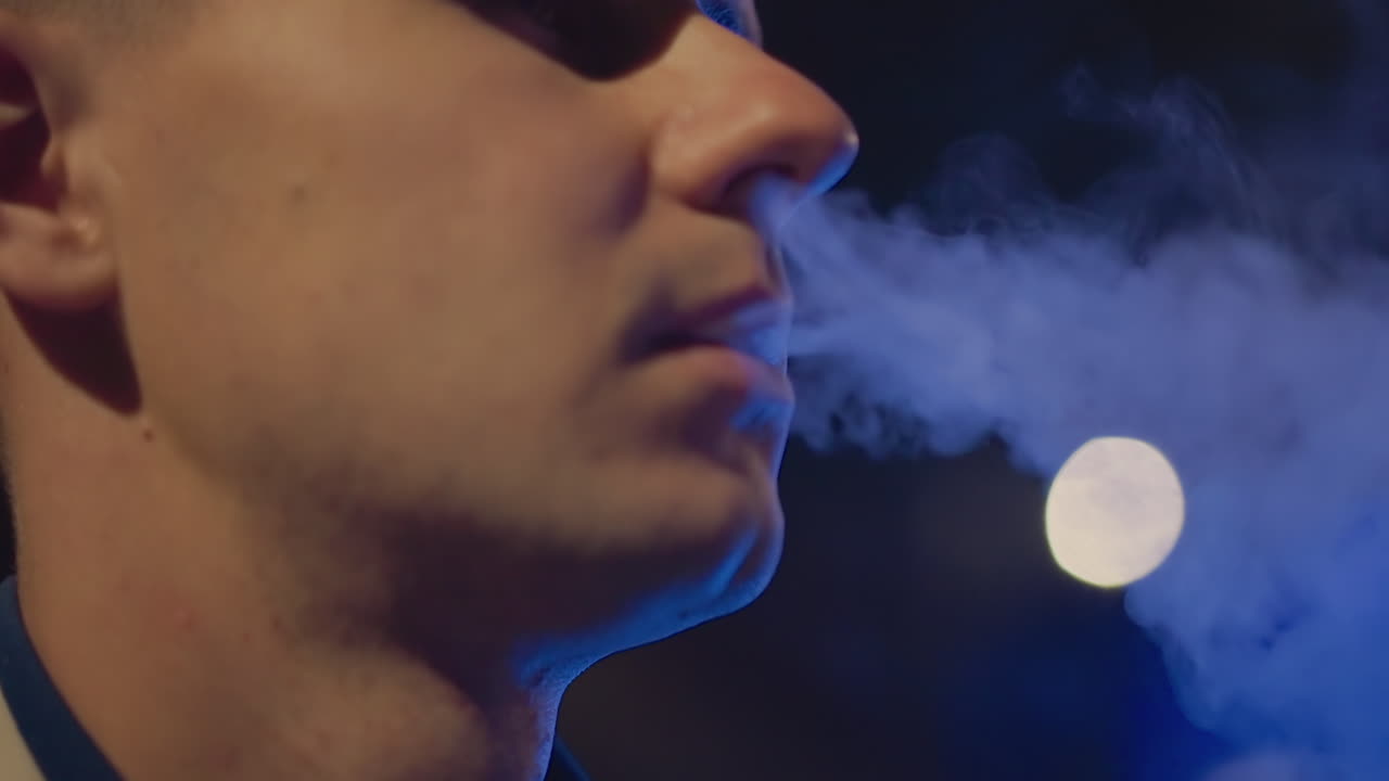 밤, 클로즈업 인물 사진, 흰 피부의 남자가 턱선을 드러내며 전자담배를 피우고 있어요. 타이트한 프레임, 부드러운 보케 조명, 제어된 흡입과 내뱉기, 컬링되는 증기 구름, 짧게 자란 수염의 질감, 친밀한 영화적 분위기, 야간
