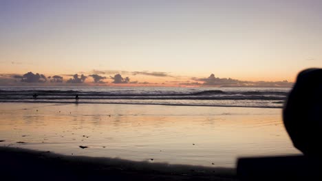 Eine-Kamera-Ist-Auf-Einem-Stativ-Aufgestellt,-Um-Bei-Ebbe-Am-Laguna-Beach-Den-Atemberaubenden-Sonnenuntergang-Zu-Fotografieren,-Während-Surfer-Surfen-Und-Die-Sonne-Zwischen-Den-Felsen-Ihren-Höhepunkt-Erreicht-Und-Sich-Im-Wasser-Spiegelt