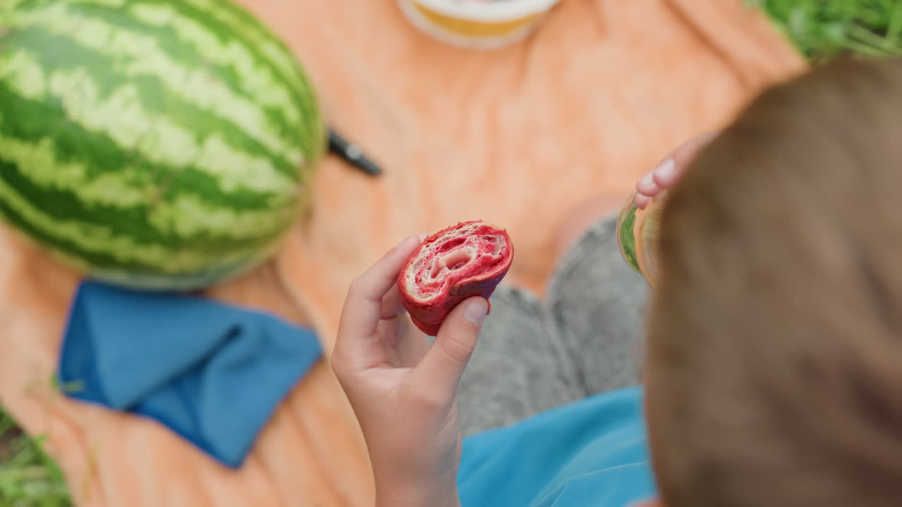 담요 위에서 페이스트리를 들고 있는 백인 소년. 수박과 토마토 그릇이 있는 피크닉 구도, 소년은 주스를 마시고, 햇빛이 비치는 풀밭, 바삭한 페이스트리와 잘 익은 멜론 조각, 편안한 공원 분위기, 캐주얼