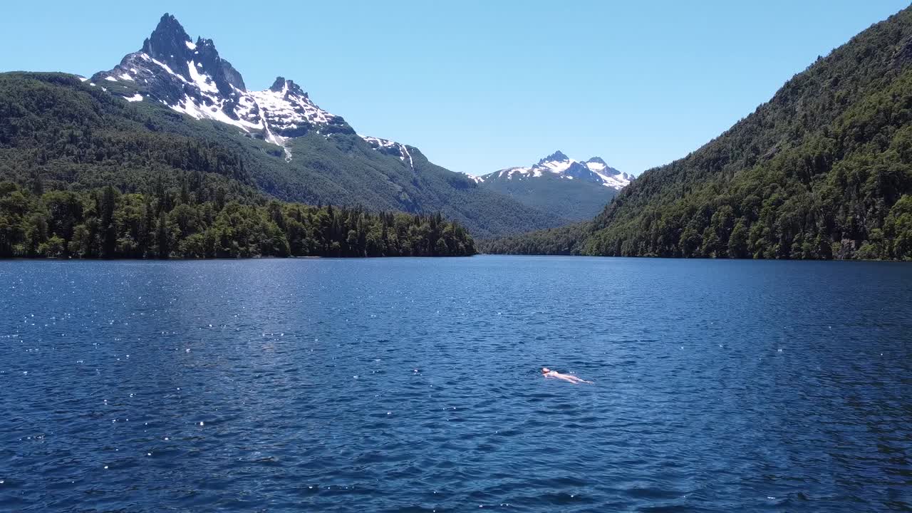 woman floating in quiet lake surrounded by green forest and snowy mountains