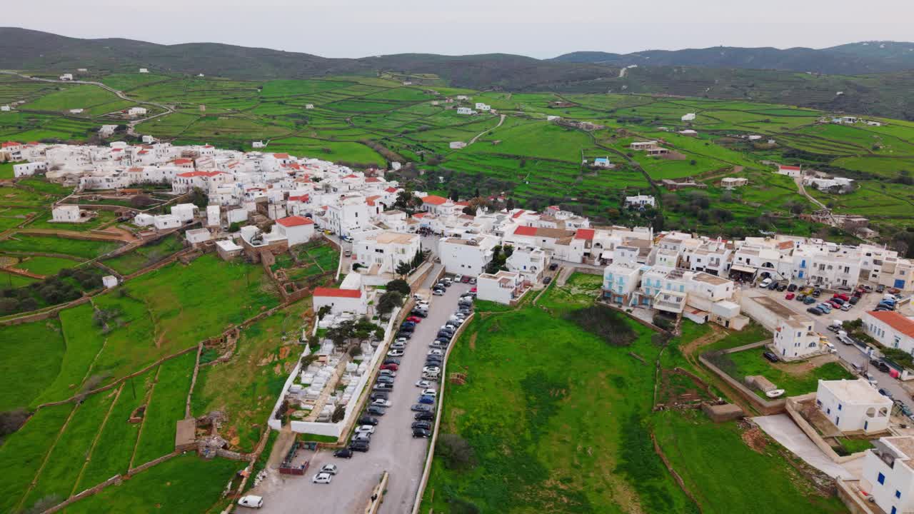 Wide aerial tracking left across of Kythnos hillside town with stone paths and terraced green backdrop, whitewashed homes
