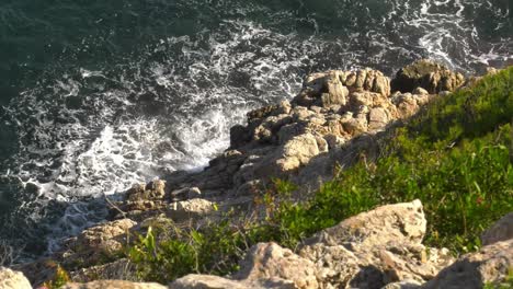 Blue-sea-breaking-on-rugged-cliffs-in-slow-motion,-birds-eye-view