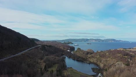 Ferry-aproaching-Sandvikvag-ferry-pier-in-the-distant---Norway-coastal-road-E39-to-ferry-pier-to-the-left-in-frame---Static-aerial-with-blue-sky-background-and-small-lake-Pollen-in-foreground