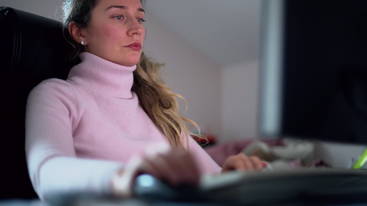 A woman sits attentively at her home office desk, working on a computer. The soft glow of the screen illuminates her focused expression as evening settles outside