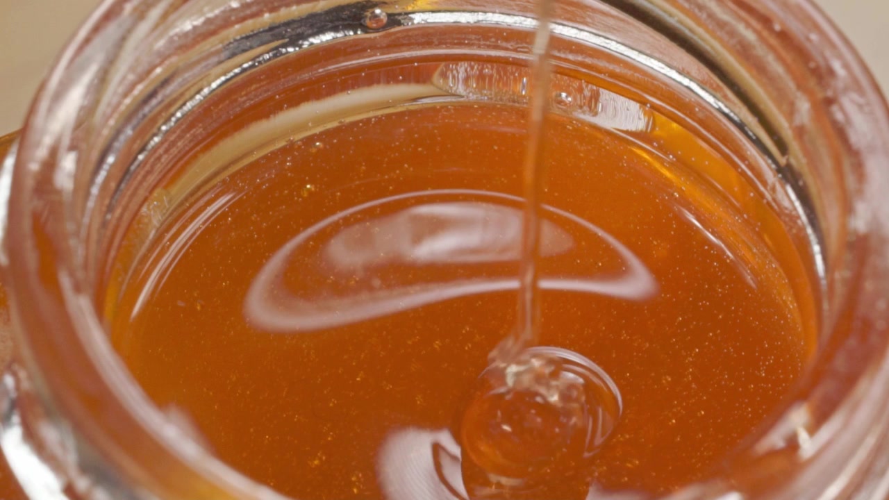 Vista macro del miele che viene versato in un barattolo di vetro, creando increspature sulla superficie dorata. Il miele denso scorre in un flusso uniforme con riflessi lucidi visibili