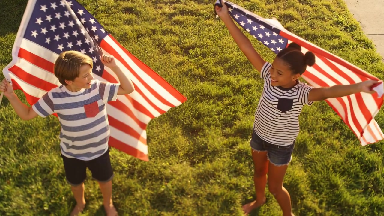 Waving two kids responding to overhead camera recording in front yard, posing with large US flags