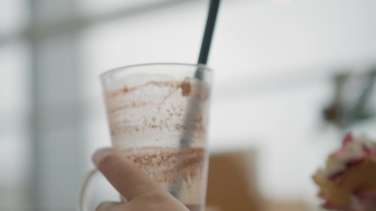 Una mano afferra una tazza di vetro trasparente con una bevanda al cioccolato schiumosa, una cannuccia inserita, un vortice di schiuma e striature di residui, la tazza leggermente inclinata durante un rinfresco casuale al chiuso in una luce diurna soffusa.
