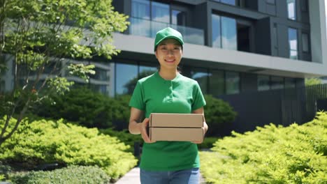 a cheerful delivery person in green attire holding a package stands outside a modern building surrounded by lush greenery, ready to make a delivery to a satisfied customer