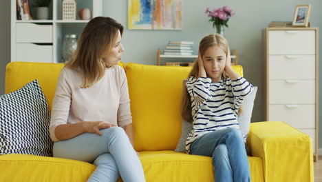 blonde mother sitting with her teenage daughter on yellow sofa and quarreling at her, girl doesn't listen and covers her ears