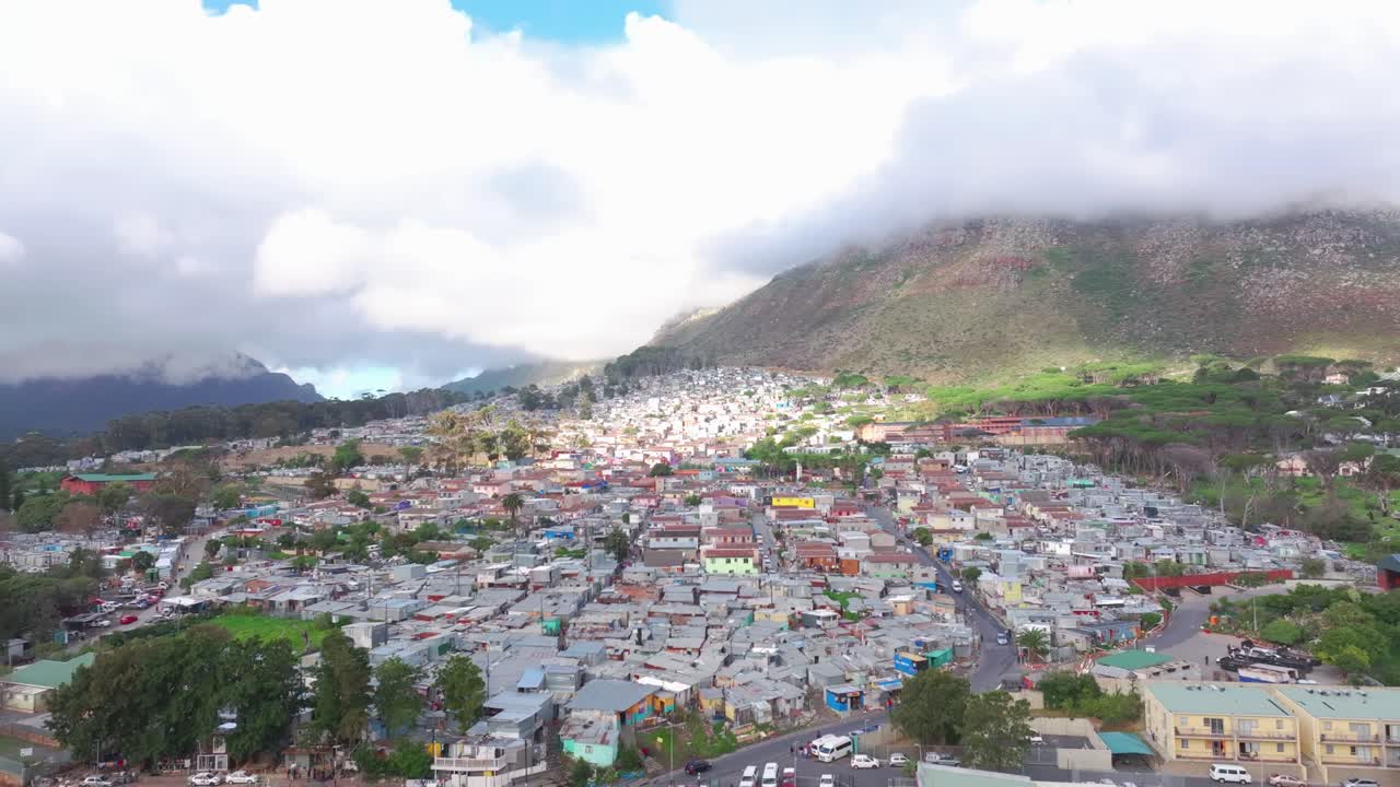 Filmato aereo in 4K con drone della township di Imizamo Yethu a Hout Bay, Città del Capo, Sudafrica. Ampia veduta panoramica che mostra un insediamento informale su uno sfondo montuoso con cielo parzialmente nuvoloso.
